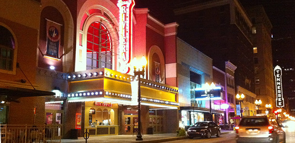 Nighttime view of historic-style theater with large lighted marquee and neon vertical sign.